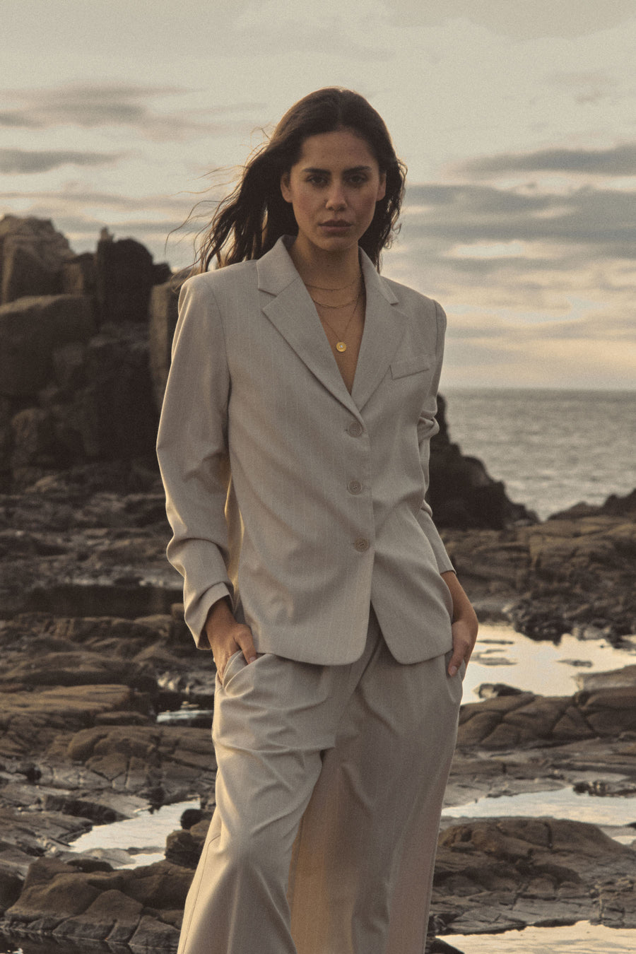 Zoe Relaxed Pinstripe Blazer in beige pinstripe worn with matching trousers on a rocky coastline